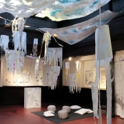 Lichte Kunstinstallation in einem holzgetäfelten Raum: Weiße Vliesbahnen und quallenartige Papierobjekte hängen über einer gemütlichen Sitzecke mit grauen Kissen.