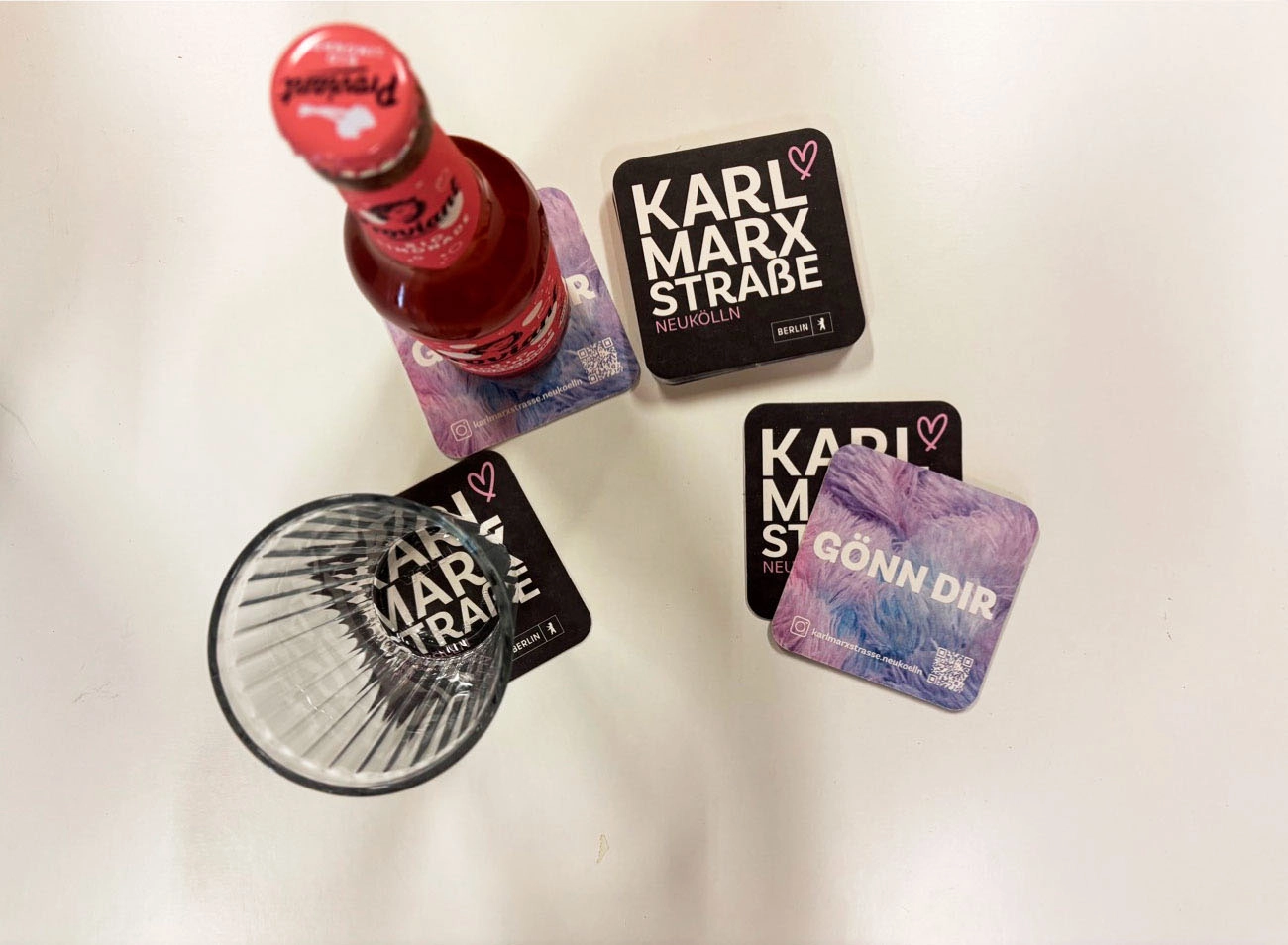 Getränk und Glas auf Untersetzern mit „Karl-Marx-Straße“- und „Gönn Dir“-Motiven.