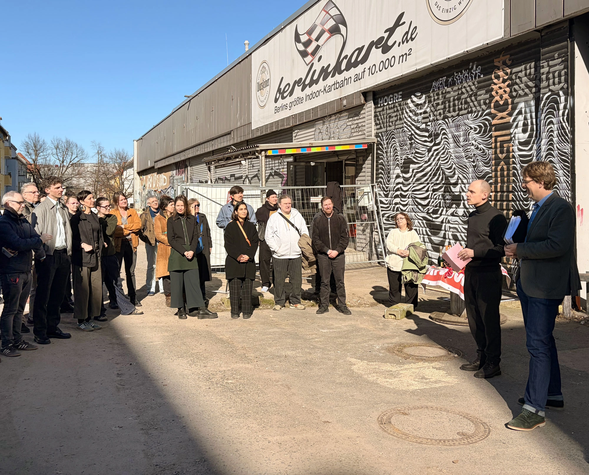 Bezirksstadtrat Jochen Biedermann auf dem Kindl-Areal erteilt die Baugenehmigung für das Großprojekt an den Vorstand der Vollgut eG. Zahlreiche Genossenschaftsmitglieder, Unterstützende, Menschen aus der Nachbarschaft und Interessierte verfolgen diesen Moment.