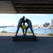 Zwei Skulpturen unter der Brücke halten ein gelbes Element am Wasser vor der Berliner Skyline, bei den Kunstaktionen auf dem Karl-Marx-Platz in Berlin.