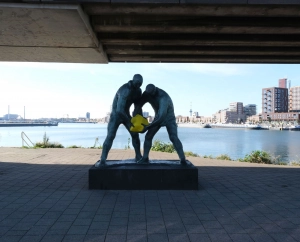 Zwei Skulpturen unter der Brücke halten ein gelbes Element am Wasser vor der Berliner Skyline, bei den Kunstaktionen auf dem Karl-Marx-Platz in Berlin.