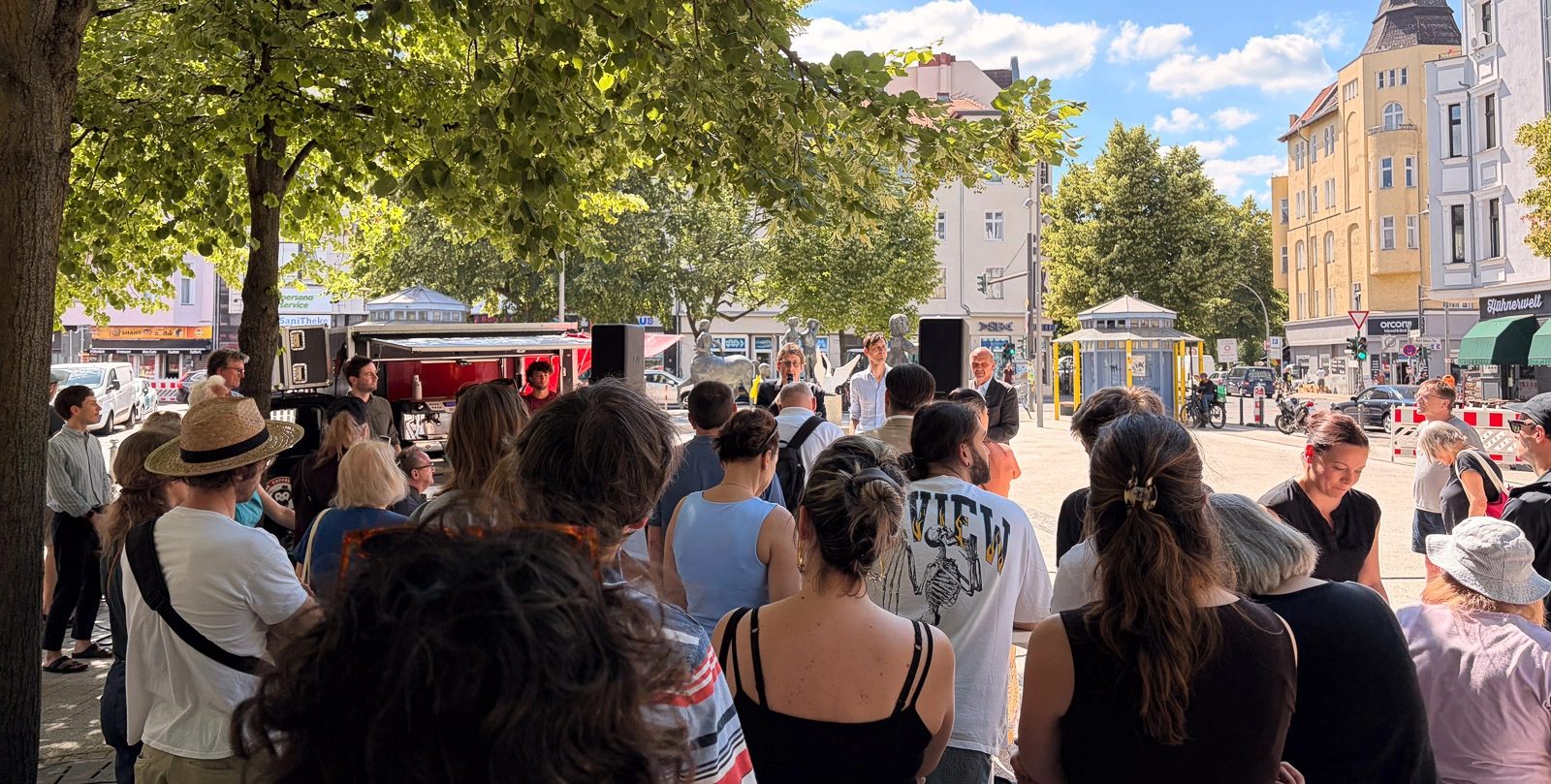 IMG_4709 Menschen versammeln sich auf dem Karl-Marx-Platz in Neukölln vor einer kleinen Bühne unter Bäumen.