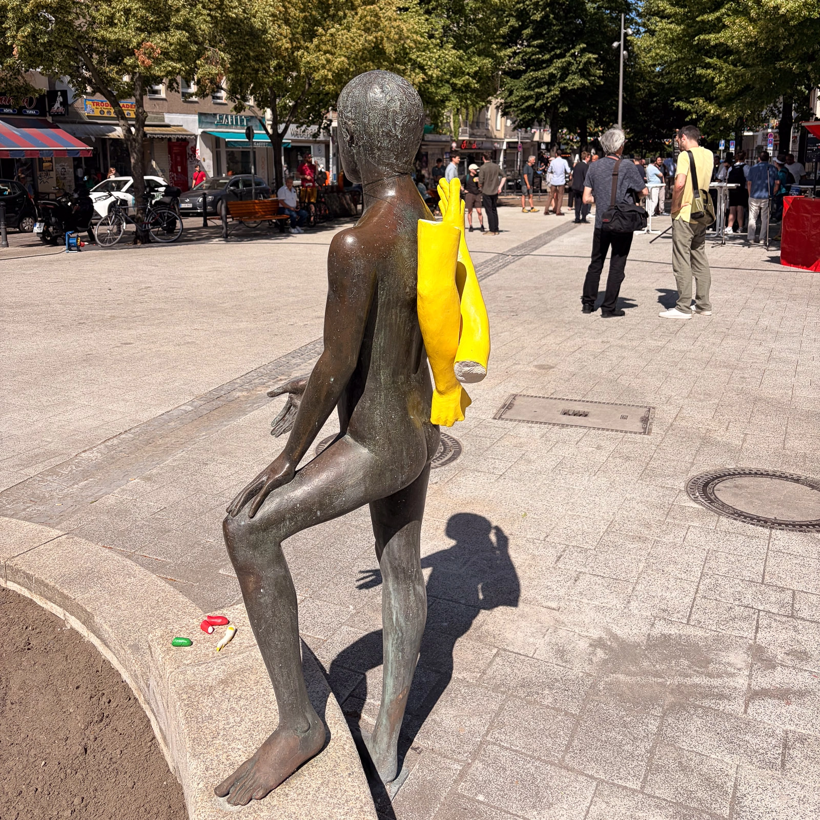 IMG_4744 Bronzefigur mit gelben Armen steht auf dem Karl-Marx-Platz, während Passanten im Schatten der Bäume vorbeigehen.