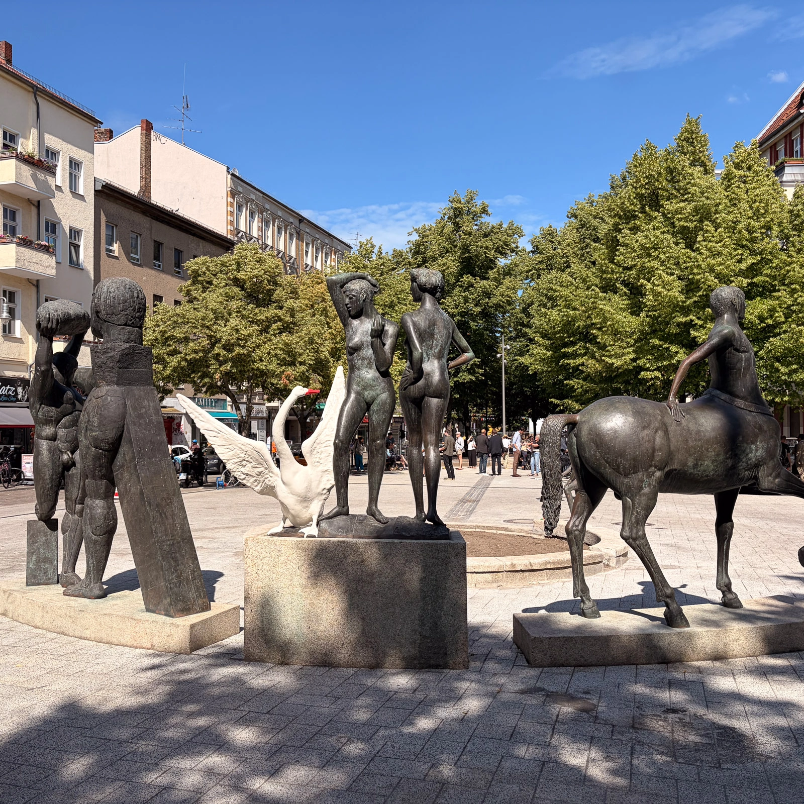 IMG_4754 Bronzeskulpturen mit menschlichen Figuren und einem Pferd auf dem Karl-Marx-Platz, umgeben von Bäumen und Häusern.