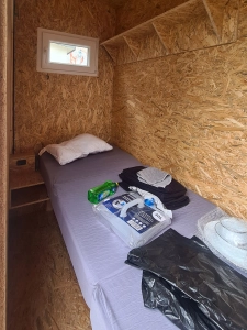 kms-sonne_20240301_123424 Kleine Holzverkleidete Kammer mit schmalem Bett, Fenster und Vorräten wie Decke, Flasche und Wasserkanister.