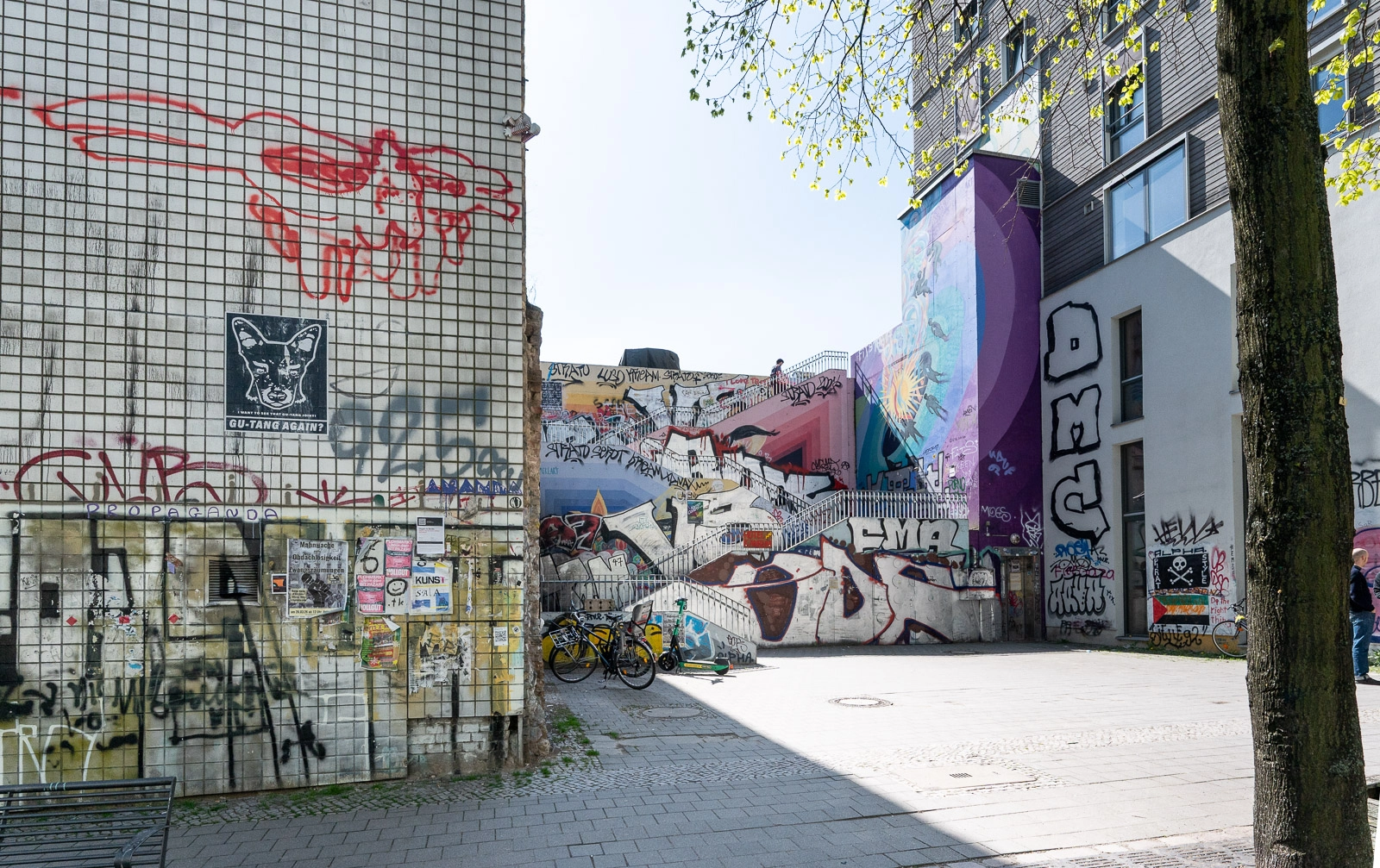Graffiti und Wandbilder bedecken eine Hofecke mit Treppen, Fahrrädern und einem hellen Platz in Berlin-Neukölln.