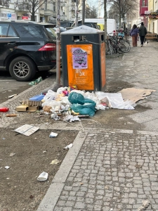 kms-sonne_IMG_1603 An der Karl-Marx-Straße in Neukölln liegt Müll um einen festgeketteten Mülleimer, während Passanten vorbeigehen.