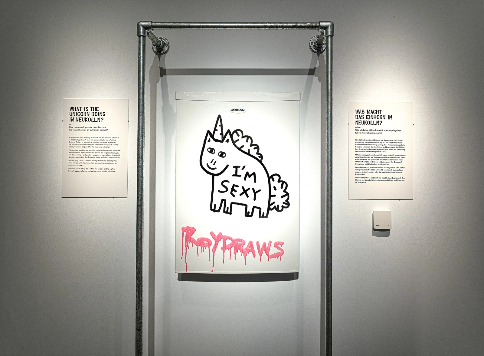 Eine Kühlschranktür mit Unicorn-Zeichnung und dem Schriftzug „I’m sexy“, flankiert von Ausstellungstexten in Neukölln.