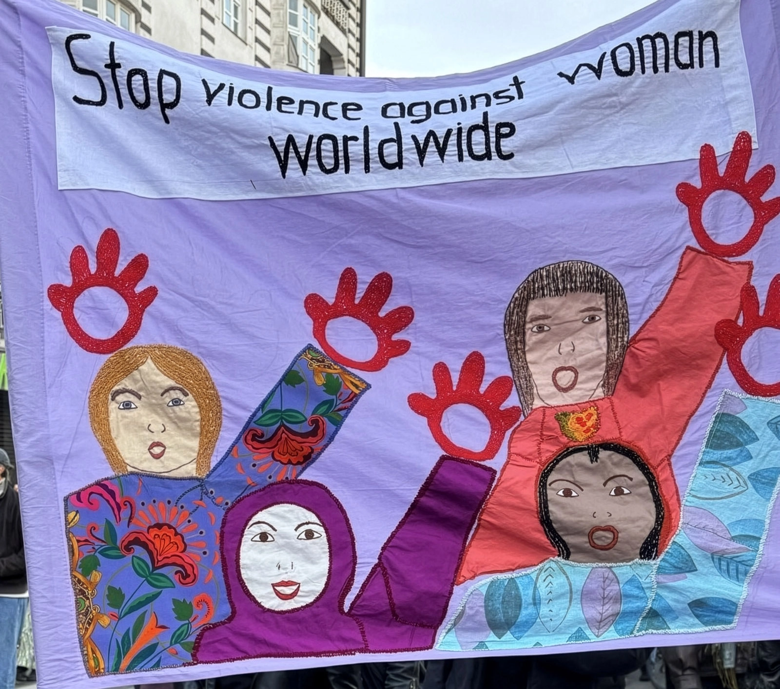 kms-sonne_imag-frauendemo Lila Demonstrationsbanner mit der Aufschrift „Stop violence against woman worldwide“ und gezeichneten Frauenfiguren.