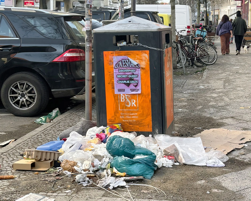 Überquellende Müllsäcke und Verpackungen liegen um einen Abfallbehälter auf einem Berliner Gehweg.