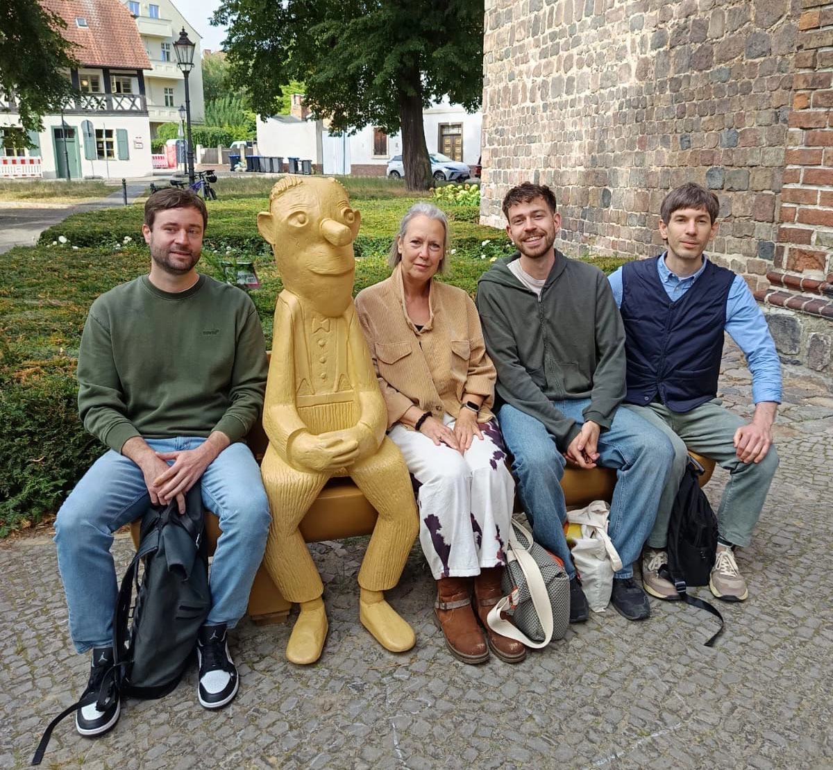 Vier Personen sitzen neben einer gelben Holzfigur auf einer Bank vor einer historischen Mauer in einem begrünten Stadtraum.